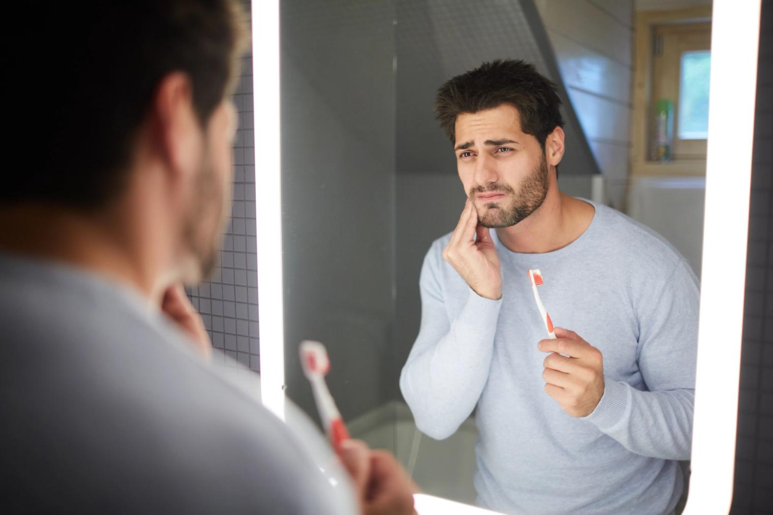 A man holding a toothbrush looks at his reflection in a mirror, clutching his jaw in discomfort.