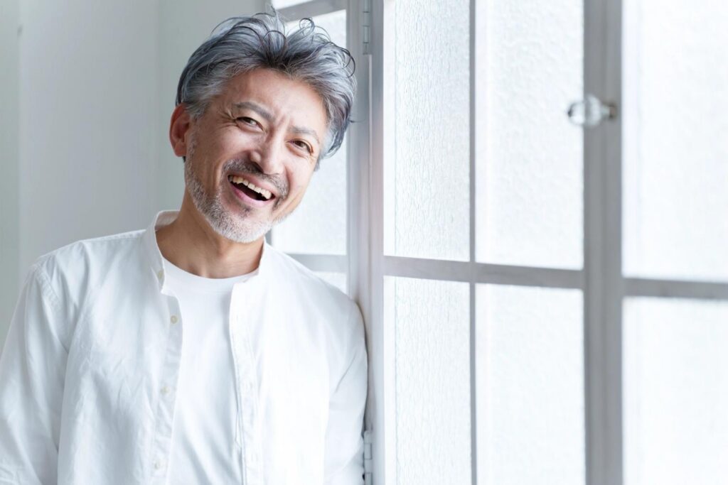 A smiling man with grey hair and a beard leans against a white window frame.