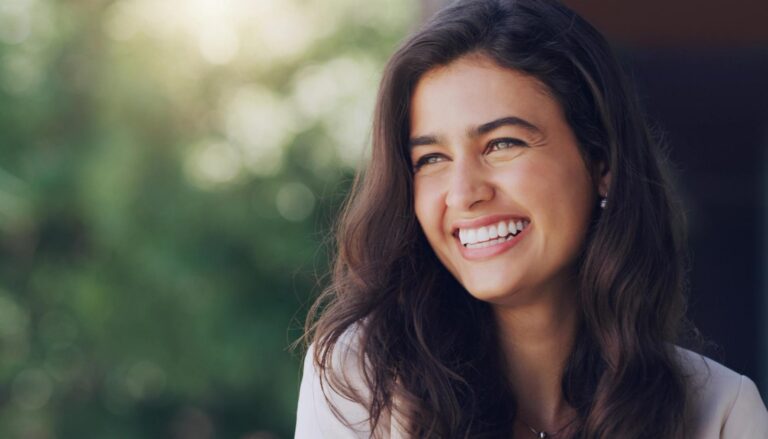A smiling woman with long, dark wavy hair looks off to the side, with a blurred green background.