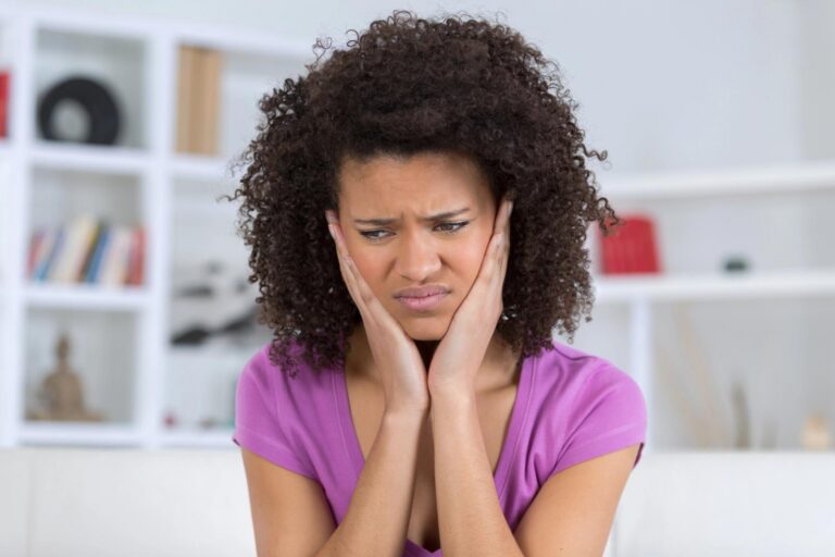 A woman with curly hair holds her cheeks with a pained expression, suggesting a toothache or jaw pain.