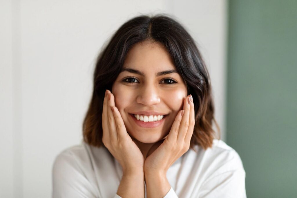 A young woman with dark hair smiling brightly with her hands on her cheeks.