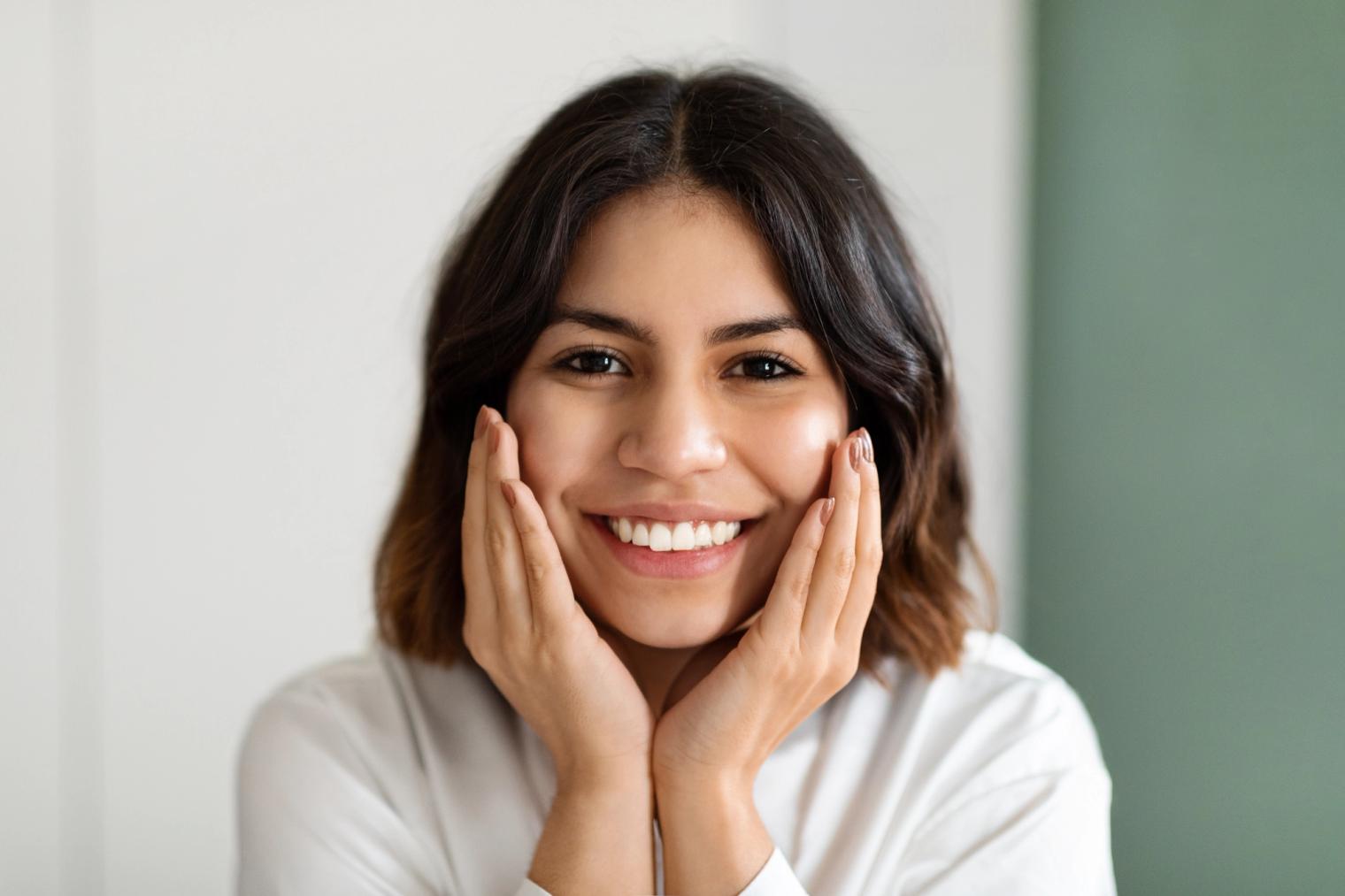 A young woman with dark hair smiling brightly with her hands on her cheeks.