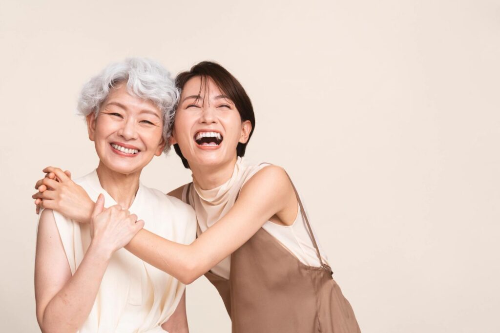 An older woman with silver hair and a younger woman with dark hair embrace and laugh joyfully.