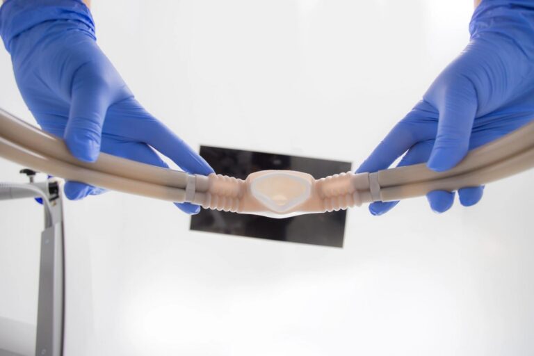 Hands in blue gloves hold a medical tube with a translucent central connector.