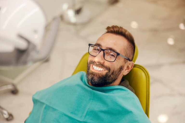 Man with glasses and beard smiling brightly in a dental chair, showing white teeth.