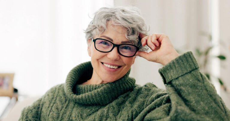 Smiling older woman with short, curly gray hair, black glasses, and a green knitted sweater.
