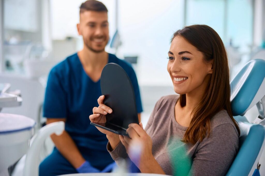 Smiling woman in a dental chair looks into a handheld mirror, with a male dentist smiling behind her.