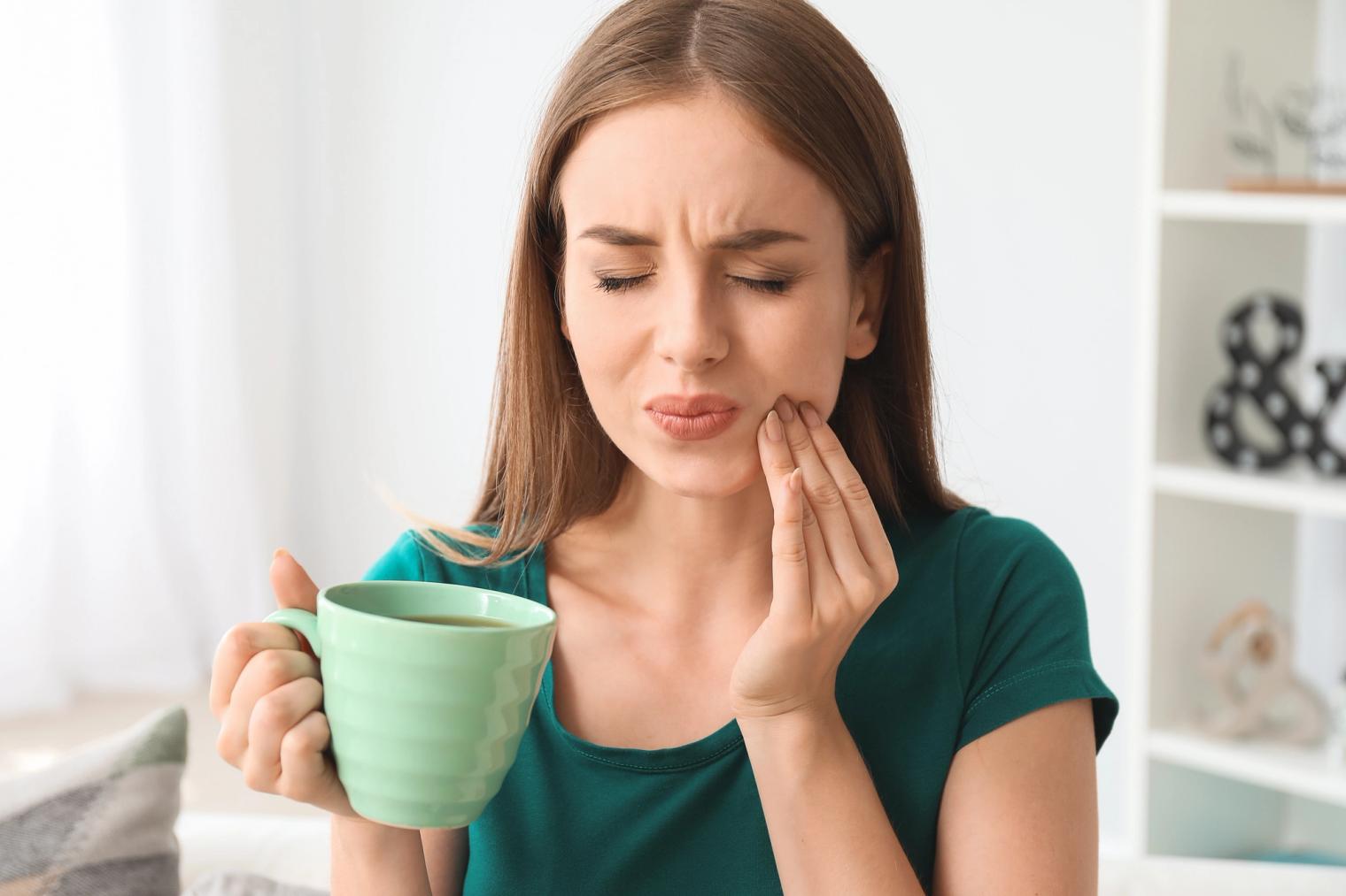 Woman grimacing and clutching her jaw in pain while holding a mug.